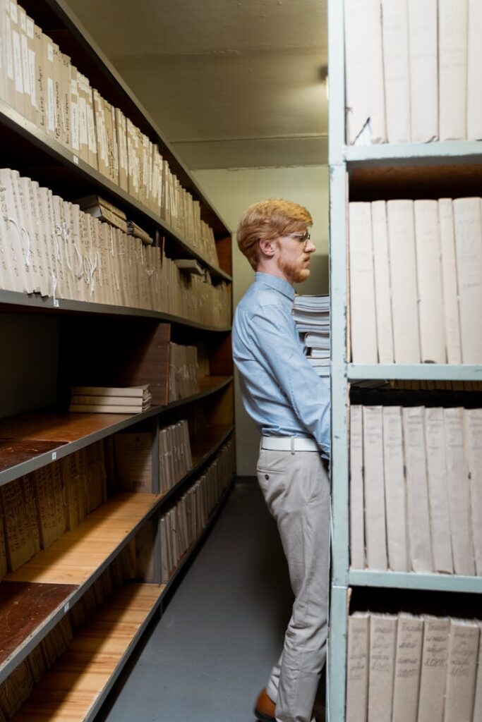 Side view of a man organizing documents in a dimly lit archive room with shelves of files.